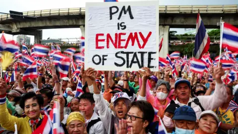 Reuters A woman standing in the centre of a crowd that fills the image, holding a placard that reads "PM is enemy of state" in black lettering, with enemy written in red. The entire crowd is filled with people waving the Thai flag. In the background is an overpass.