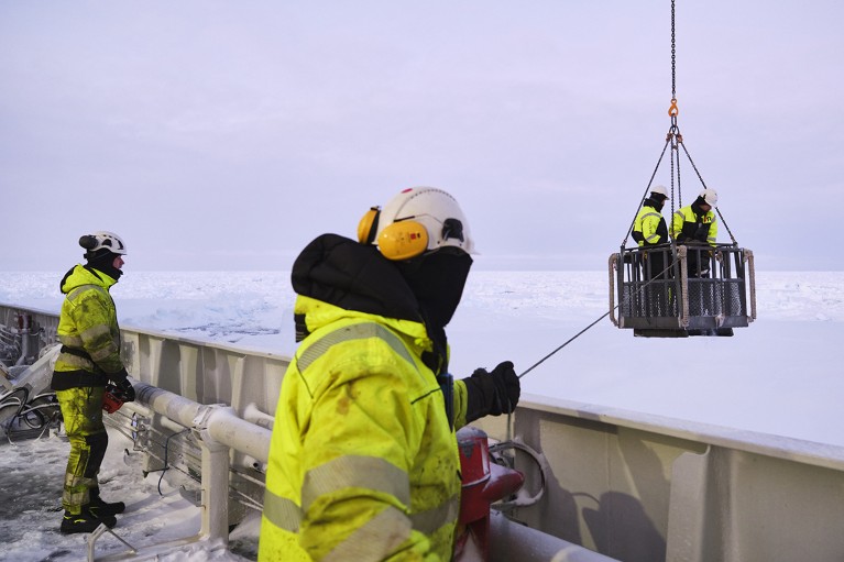 Two crew members on a ship deploy two scientists with a wave buoy for an ice floe in the Greenland sea to study sea ice dynamics.