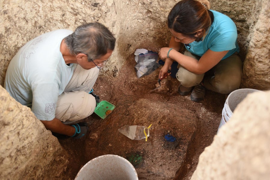 A man and woman crouch in an earthen pit looking at the ground where bright blue and green patches of color are visible