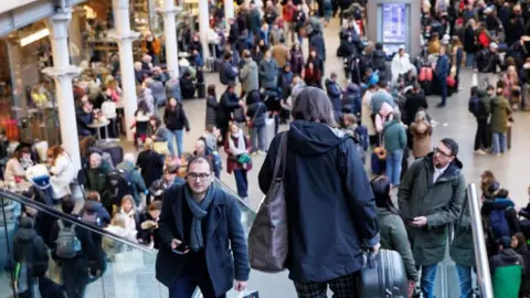 EPA A woman holding a suitcase, dressed in a navy blue jacket with a large handbag slung over her shoulder, makes her way down the stairs at a train station, with crowds of people gathered on the platform below. 