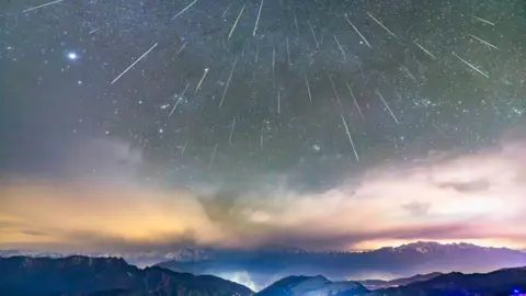 Getty Images Multiple bright meteor streaks from the Geminid meteor shower radiate across a star-filled sky above layered mountain ranges, with illuminated roads and lights visible on the slopes of Niubei Mountain in Ya’an, Sichuan Province, China.