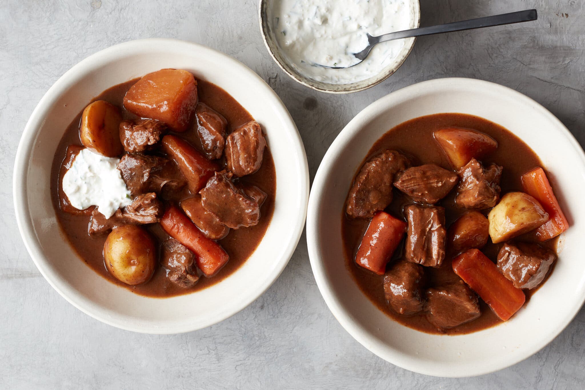 Identical white bowls hold beef stew with large carrot and potato chunks. A small dish of herb-flecked sour cream is visible in the background.